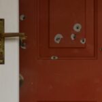 Red front door with multiple bullet holes and a brass handle on the left side, close-up view.