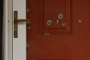Red front door with multiple bullet holes and a brass handle on the left side, close-up view.