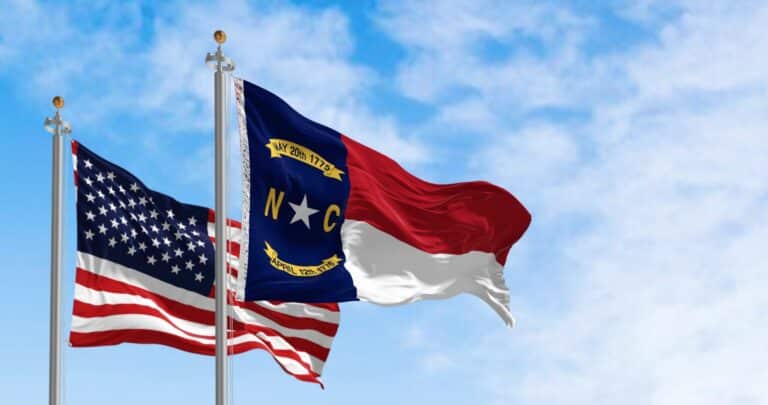 Two flags—United States and North Carolina state flag—flying on separate flagpoles against a bright blue sky with clouds.