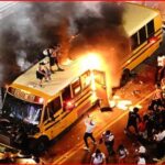 Yellow school bus on a city street engulfed in flames with dark smoke rising, surrounded by bystanders and onlookers at nightfall.