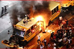 Yellow school bus on a city street engulfed in flames with dark smoke rising, surrounded by bystanders and onlookers at nightfall.