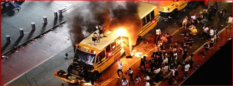 Yellow school bus on a city street engulfed in flames with dark smoke rising, surrounded by bystanders and onlookers at nightfall.