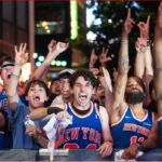 Crowd of basketball fans in blue–orange New York jerseys cheering with raised arms behind a barrier.