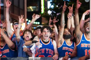 Crowd of basketball fans in blue–orange New York jerseys cheering with raised arms behind a barrier.