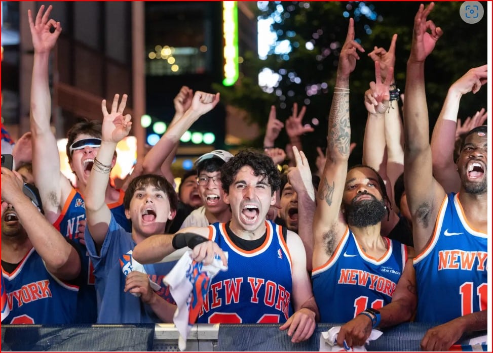 Crowd of basketball fans in blue–orange New York jerseys cheering with raised arms behind a barrier.