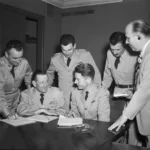 Five military officers in dress uniforms gather around a table as two sign documents; a suited man observes nearby in a formal briefing.
