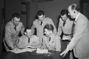 Five military officers in dress uniforms gather around a table as two sign documents; a suited man observes nearby in a formal briefing.