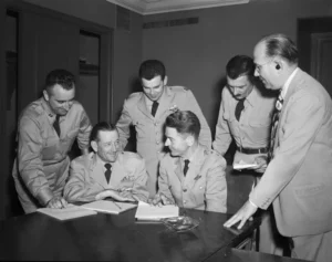 Five military officers in dress uniforms gather around a table as two sign documents; a suited man observes nearby in a formal briefing.