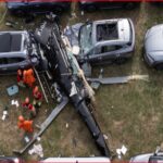 A damaged black helicopter lies between parked cars in a grassy lot with rescue workers in orange uniforms nearby.