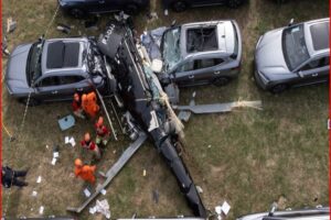 A damaged black helicopter lies between parked cars in a grassy lot with rescue workers in orange uniforms nearby.