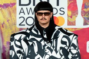 Person wearing a black wig, sunglasses, and a bold black-and-white patterned suit posing at an awards backdrop.