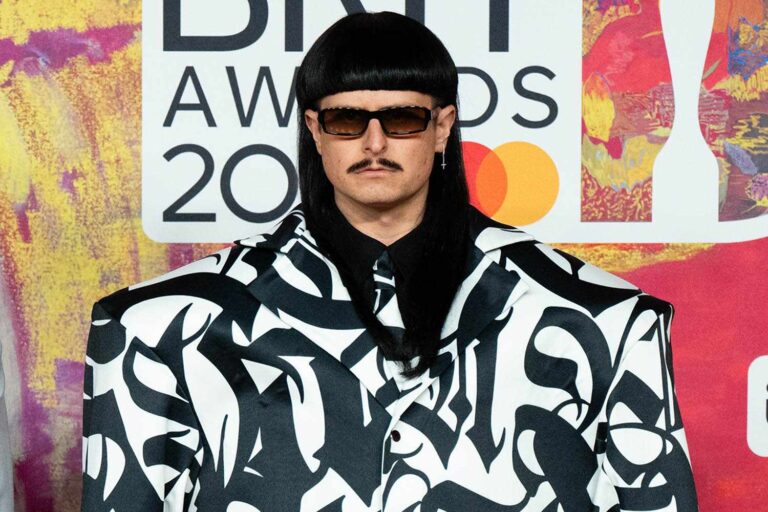 Person wearing a black wig, sunglasses, and a bold black-and-white patterned suit posing at an awards backdrop.