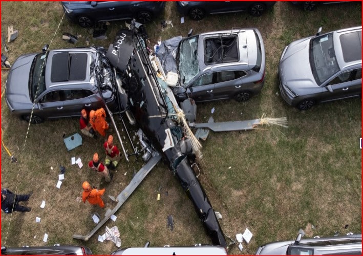 A damaged black helicopter lies between parked cars in a grassy lot with rescue workers in orange uniforms nearby.