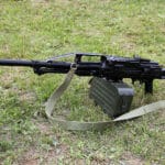 Black rifle with bipod and a green ammo box resting on a grassy field with a tan strap nearby.