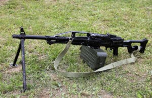 Black rifle with bipod and a green ammo box resting on a grassy field with a tan strap nearby.