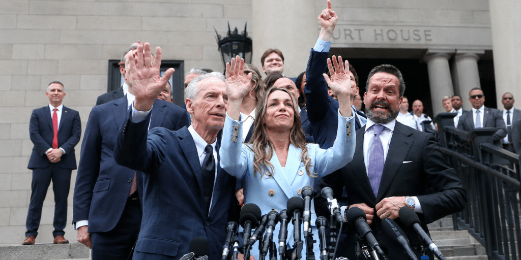 Politicians on courthouse steps raise their hands in a victory gesture, lined with microphones in the foreground, a press conference moment.