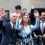 Politicians on courthouse steps raise their hands in a victory gesture, lined with microphones in the foreground, a press conference moment.