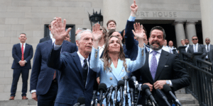 Politicians on courthouse steps raise their hands in a victory gesture, lined with microphones in the foreground, a press conference moment.
