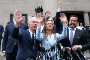 Politicians on courthouse steps raise their hands in a victory gesture, lined with microphones in the foreground, a press conference moment.