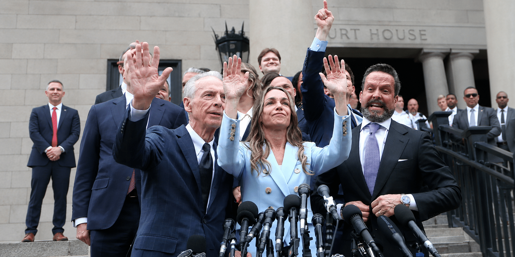 Politicians on courthouse steps raise their hands in a victory gesture, lined with microphones in the foreground, a press conference moment.