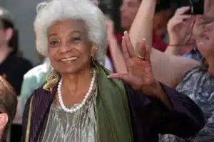 Smiling elderly woman with white hair wearing a pearl necklace waves to a crowd at an event