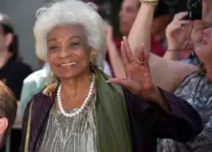 Smiling elderly woman with white hair wearing a pearl necklace waves to a crowd at an event