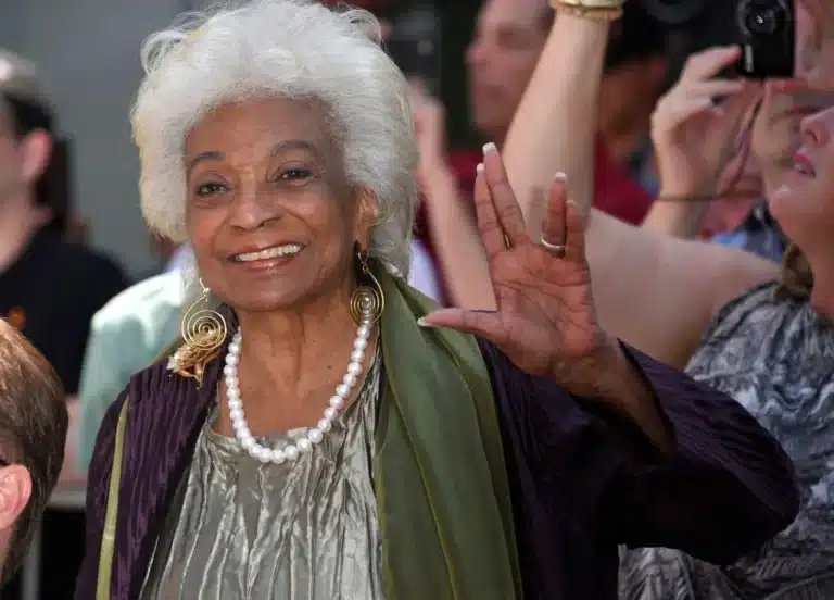 Smiling elderly woman with white hair wearing a pearl necklace waves to a crowd at an event