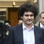 Curly-haired man in a navy suit escorted by armed police outside a building.