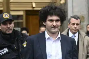 Curly-haired man in a navy suit escorted by armed police outside a building.