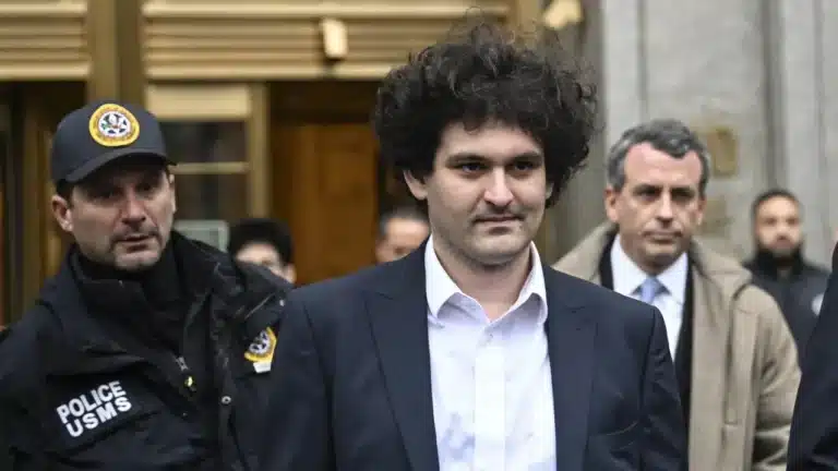 Curly-haired man in a navy suit escorted by armed police outside a building.