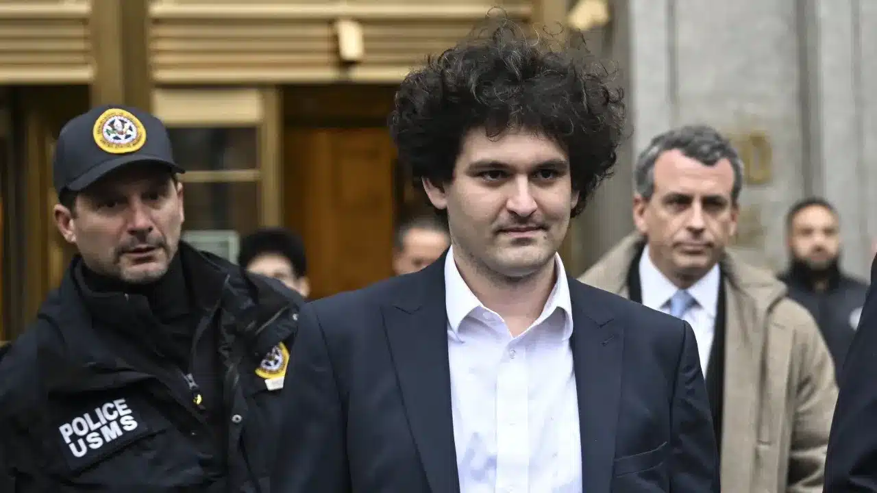 Curly-haired man in a navy suit escorted by armed police outside a building.
