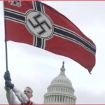 Person raises a large Nazi swastika flag in front of the U.S. Capitol under a gray sky, with a demonstrator visible nearby.