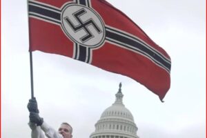 Person raises a large Nazi swastika flag in front of the U.S. Capitol under a gray sky, with a demonstrator visible nearby.