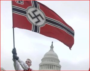 Person raises a large Nazi swastika flag in front of the U.S. Capitol under a gray sky, with a demonstrator visible nearby.