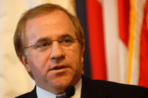 Middle-aged man in a suit and glasses speaks into a microphone, with red and flag banners in the background.