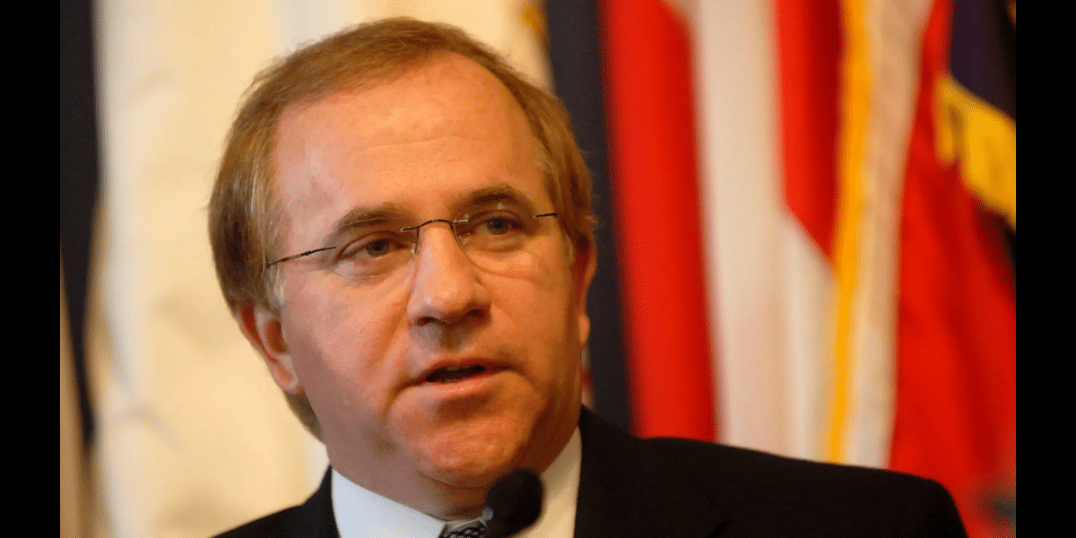 Middle-aged man in a suit and glasses speaks into a microphone, with red and flag banners in the background.