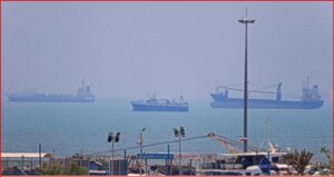 Harbor scene with cranes and warehouses in the foreground and several large cargo ships on a calm, hazy sea in the distance.