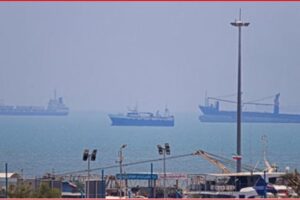 Harbor scene with cranes and warehouses in the foreground and several large cargo ships on a calm, hazy sea in the distance.