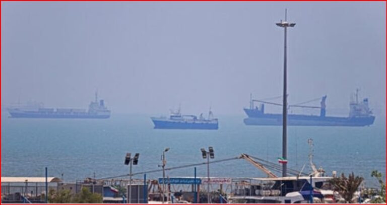 Harbor scene with cranes and warehouses in the foreground and several large cargo ships on a calm, hazy sea in the distance.