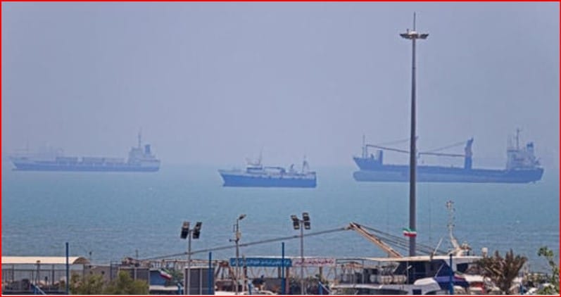 Harbor scene with cranes and warehouses in the foreground and several large cargo ships on a calm, hazy sea in the distance.