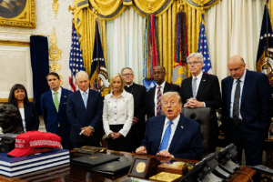 President Donald Trump seated at a desk signing papers as aides stand behind in a gold ornate room with flags and framed documents, a red MAGA hat on the desk.
