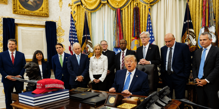 President Donald Trump seated at a desk signing papers as aides stand behind in a gold ornate room with flags and framed documents, a red MAGA hat on the desk.
