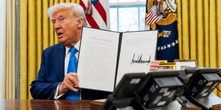 Man in a blue suit holds up an open, signed document in a formal office with gold curtains and flags in the background.