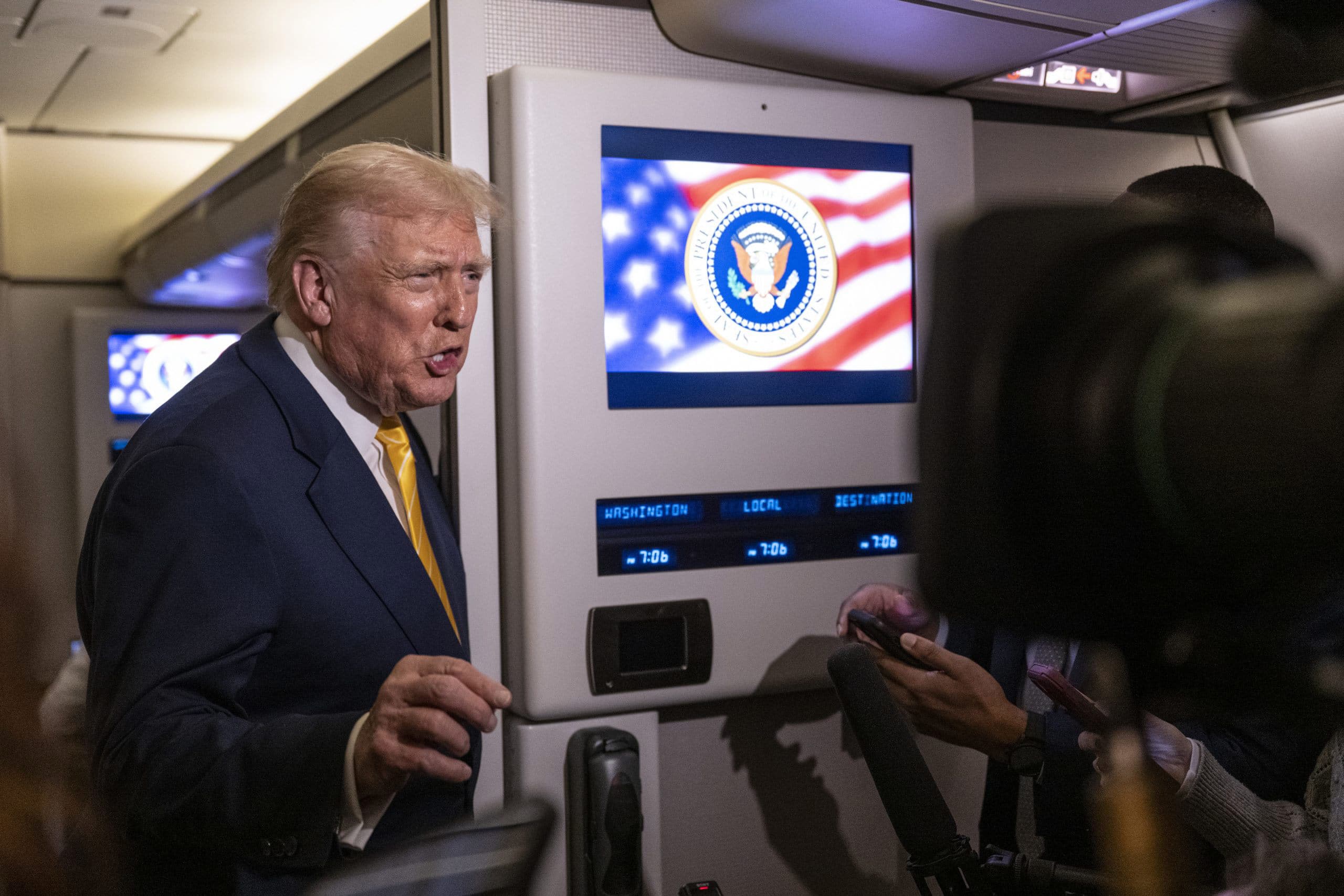 Former President Donald Trump speaks to reporters on an airplane, with a screen showing the presidential seal behind him.