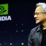 Side profile of a man in a black leather jacket speaking on stage with a large NVIDIA logo in the green-lit background to the left.