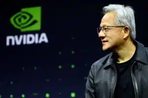 Side profile of a man in a black leather jacket speaking on stage with a large NVIDIA logo in the green-lit background to the left.