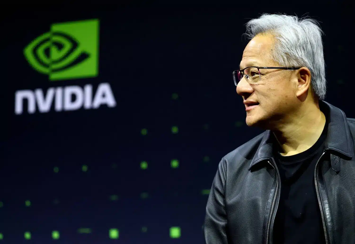 Side profile of a man in a black leather jacket speaking on stage with a large NVIDIA logo in the green-lit background to the left.