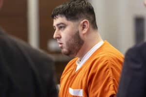 Profile of a man in an orange jail jumpsuit, beige walls in the background, looking down thoughtfully.