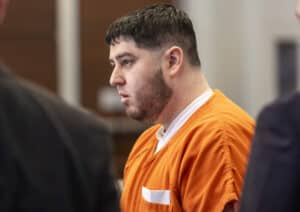 Profile of a man in an orange jail jumpsuit, beige walls in the background, looking down thoughtfully.