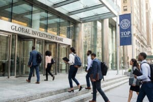 People in business attire entering a glass-front Global Investment Bank entrance; a 'Top University' banner is visible nearby outside the building.